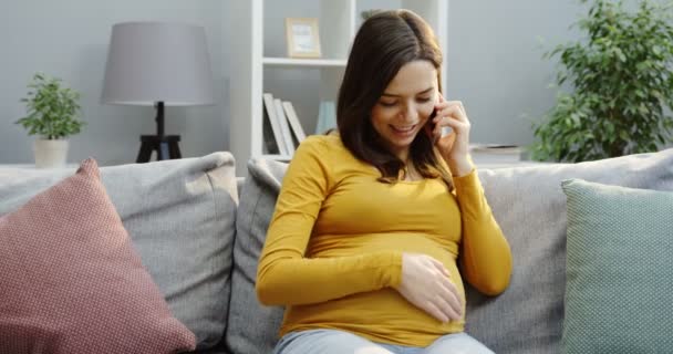Csinos mosolygós terhes nő simogatja a nagy hasát, és a kanapén beszélget a mobilján párnákkal a kényelmes fényszobában. Beltéri — Stock videók