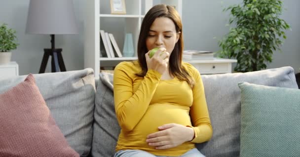 Ganska gravid kvinna som vilar på den grå soffan bland kuddar, biter och äter ett grönt äpple i det mysiga rummet hemma. Inomhus — Stockvideo