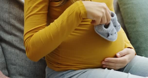 Close up of the big pregnant belly and womans hands playing with small kids sleepers near it. on the gray sofa. Inside — Stock Video