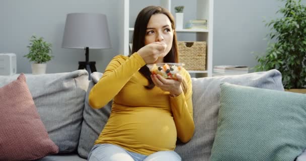 Härligt leende gravid kvinna smekte sin stora mage och äter en fruktsallad från en glasskål på soffan med kuddar i det mysiga vardagsrummet. Inuti — Stockvideo