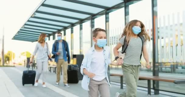 Cute small Caucasian happy kids, brother and sister in medical masks running at train station. Parents walking on blurred background and carrying suitcases on wheels. Trip concept in pandemic. — Stock Video