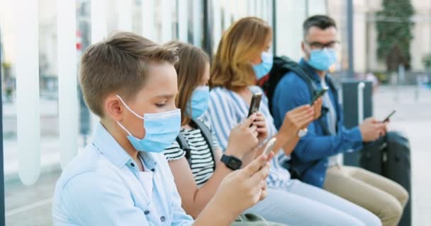 Familia caucásica con niños con máscaras médicas sentados en la parada de autobús, esperando el transporte. Niño y niña con madre y padre usando teléfonos. Niños y adultos tocando y desplazándose en teléfonos inteligentes. — Vídeos de Stock