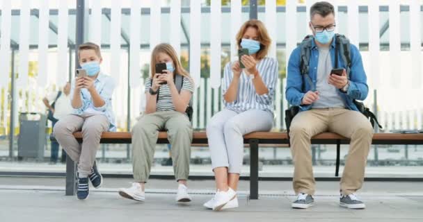 Distancia social en la parada de autobús entre los familiares. Padres y niños caucásicos con máscaras médicas sentados en el banco y usando o jugando en teléfonos móviles. Toque y desplazamiento en los teléfonos inteligentes. Viaje pandémico — Vídeo de stock