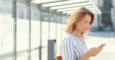 Güneşli bir günde açık havada akıllı telefon kullanan beyaz, güzel, sarışın bir kadın. Otobüs durağındaki cep telefonundan mesaj atan güzel bir kadın. Cep telefonuna bağlanan ve kayan kadın, nakil için bekliyor..