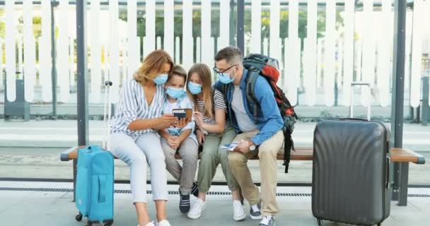 Caucasian happy family in medical masks sitting on bus stop and looking for route on tablet device. Parents with kids and suitcases on wheels waiting for transport and using gadget. Quarantine trip. — Stock Video