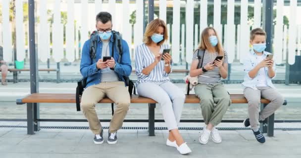 Distancia social en la parada de autobús entre los familiares. Padres y niños caucásicos con máscaras médicas sentados en el banco y usando o jugando en teléfonos móviles. Toque y desplazamiento en los teléfonos inteligentes. Viaje pandémico — Vídeo de stock