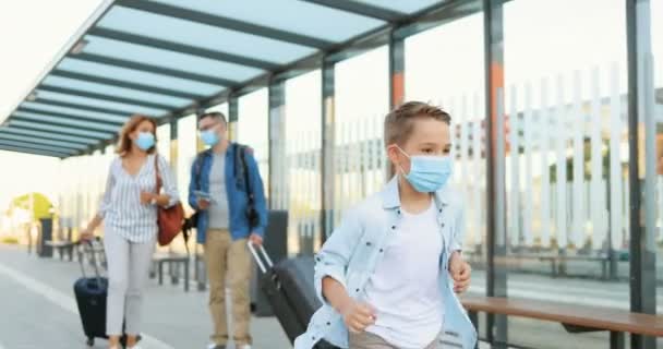 Carino piccolo ragazzo felice caucasico in maschera medica che corre alla stazione ferroviaria. Genitori che camminano su sfondo sfocato e trasportano valigie su ruote. Concetto di viaggio in pandemia. Allegro figlio correre. — Video Stock