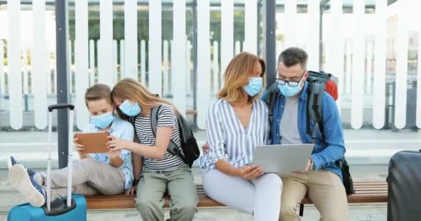 Caucasian family in medical masks sitting on bench with suitcases and waiting for bus at stop outdoor. Kids, brother and sister playing on tablet device. Parents using laptop computer for route plan. — Stock Video