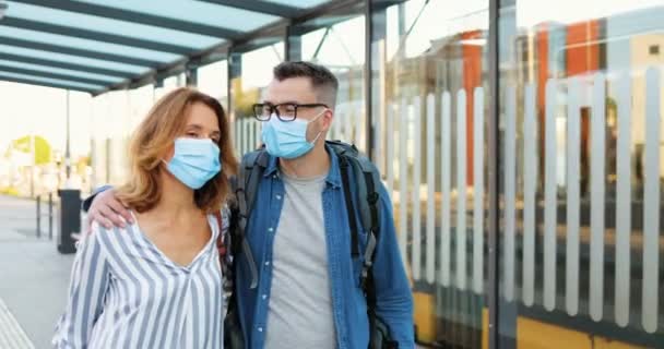 Caucasian handsome man and beautiful woman walking outdoors, talking and hugging. Couple of travellers in medical masks strolling at bus stop ot train station. Pandemic travelling. Covid-19 concept. — Stock Video