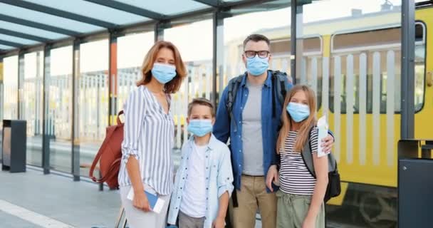 Kaukázusi boldog család portréja orvosi maszkban és bőröndökkel a buszmegállóban vagy a vasútállomáson. A szülők a fiukkal és lányukkal világjárvány idején utaznak. Közelítés. — Stock videók