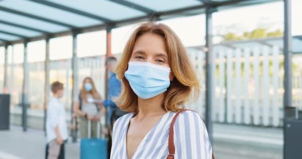 Ritratto di bella donna bionda caucasica in maschera medica guardando la macchina fotografica e in piedi alla fermata dell'autobus o alla stazione ferroviaria. Marito con bambini e valigie in attesa di trasporto su sfondo sfocato. — Video Stock