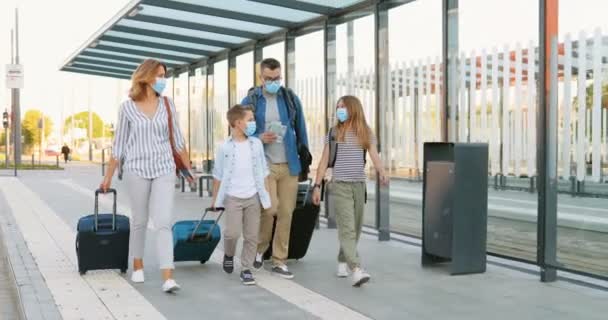 Blanke gelukkige familie met twee schattige kleine kinderen wandelen bij bushalte of treinstation, het dragen van koffers op wielen en praten. Ouders met kleine dochter en zoon in medische maskers op reis. — Stockvideo