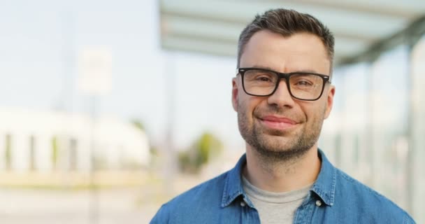 Portret van een knappe blanke man met een bril die recht en glimlachend naar de camera kijkt. Aantrekkelijk gelukkig mannetje in bril bij bushalte op zomerse zonnige dag. Sluit maar af. Inzoomen. — Stockvideo