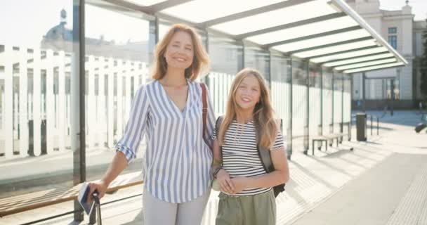 Portret młodej białej pięknej matki i uroczej nastolatki pięknej córki stojącej na przystanku autobusowym z walizką na kołach i plecakiem. słoneczny dzień. letnie wakacje. Kobieta i dziewczyna na dworcu kolejowym — Wideo stockowe