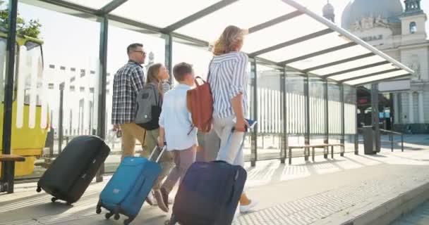 Parte trasera de la familia feliz caucásica con dos niños pequeños y lindos caminando en la parada de autobús o estación de tren, llevando maletas en ruedas y hablando. Padres con hija e hijo viajando. Vista trasera. — Vídeos de Stock