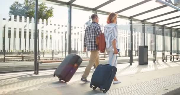Zadní na kavkazské šťastný mladý manželský pár zamilovaný chůze na autobusové zastávce a nesoucí kufry na kolech. Pohled zezadu na krásnou ženu a pohledného muže cestující a procházky na vlakovém nádraží. — Stock video