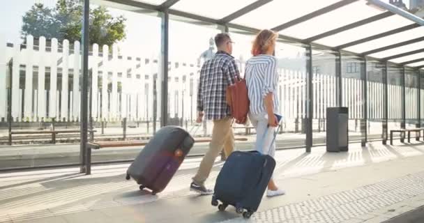 Hinten auf kaukasisch glückliches junges verliebtes Ehepaar, das an der Bushaltestelle spazieren geht und Koffer auf Rädern trägt. Rückansicht einer schönen Frau und eines gutaussehenden Mannes, die am Bahnhof unterwegs sind und spazieren gehen. — Stockvideo