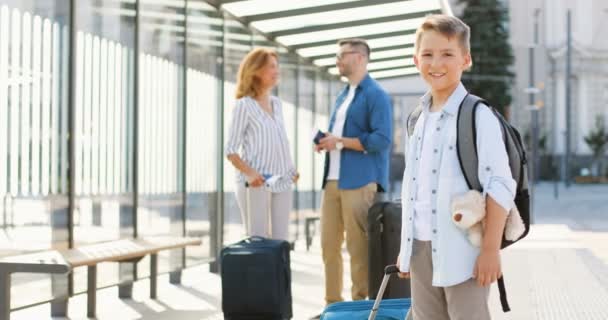 Portrait of cute Caucasian small boy with backpack, teddy bear toy and suitcase standing outdoor at bus stop and smiling to camera. Parents talking on background. Family having trip. Travelling. — Stock Video