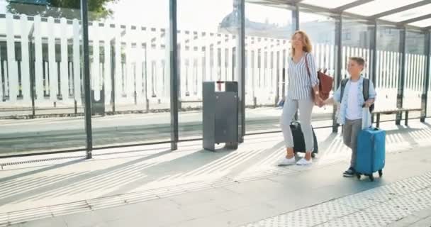 Kaukasische junge schöne glückliche Mutter und niedlicher kleiner Sohn, die die Straße entlang laufen und Koffer auf Rädern an der Bushaltestelle tragen. Frau und kleiner Junge lächeln, reden und machen Ferien. Bahnhof. — Stockvideo