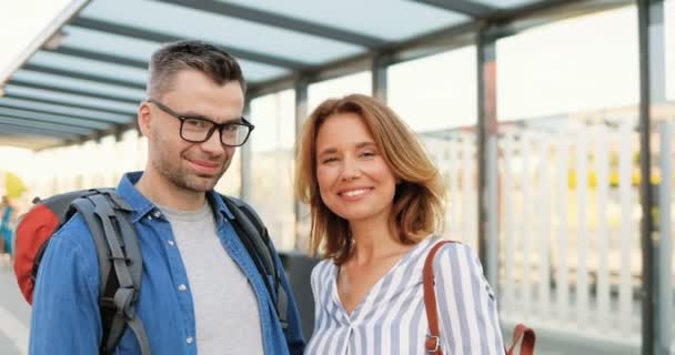 Portrait of Caucasian ceria happy couple of tourist standing at bus stop, looking at each other and smile. Wanita cantik dan pria tampan menatap kamera dengan senyum gembira di stasiun kereta api. — Stok Video