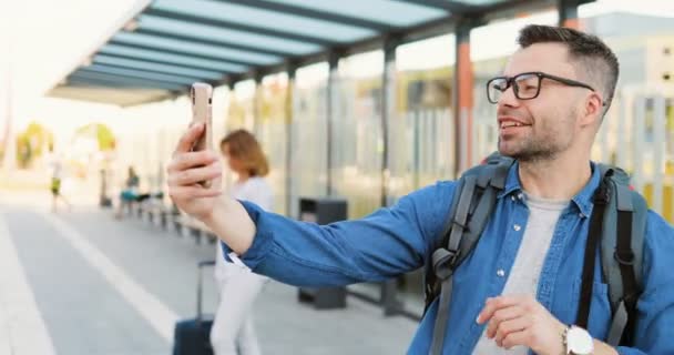 Белый красивый молодой человек в очках и с рюкзаком, стоящим на автобусной остановке и разговаривающим по мобильному телефону через веб-камеру. Мужчина в очках, разговаривающий по мобильному на вокзале. Видеочат. — стоковое видео