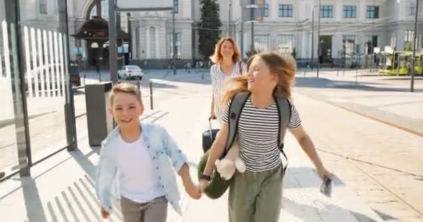 Leuke kleine blanke gelukkige kinderen, broer en zus rennend op het treinstation. Moeder loopt op de achtergrond en draagt koffers op wielen. Reisconcept. Mam volgt kinderen bij bushalte. — Stockvideo