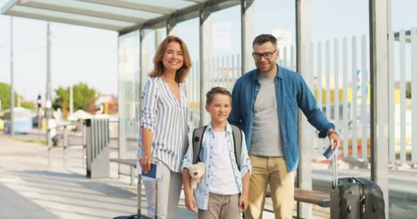 Портрет кавказской счастливой семьи с чемоданами, стоящими на улице на автобусной остановке или вокзале. Родители с сыном путешествуют летом. Отпуск ребенка с матерью и отцом Отъезд на праздники — стоковое видео