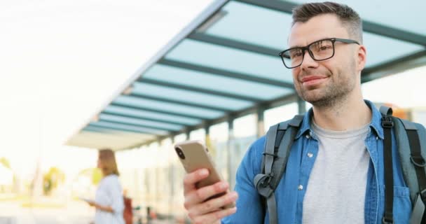 Gözlüklü, sırt çantalı, otobüs durağında duran ve cep telefonunu dinleyen beyaz tenli yakışıklı bir genç. Gözlüklü bir erkek tren istasyonunda cep telefonundan mesaj yolluyor.. — Stok video