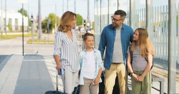 Retrato de la familia caucásica feliz con maletas de pie al aire libre en el autobús sop o estación de tren. Padres con hijo e hija viajando en verano. Vacaciones de niños con madre y padre. — Vídeo de stock