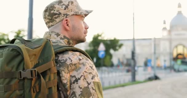 Partea din spate a unui tânăr soldat caucazian în uniformă, pălărie și cu rucsac care merge în aer liber. Vedere din spate pe ofițerul militar plimbându-se pe stradă în haine militare. Mergând în armată . — Videoclip de stoc