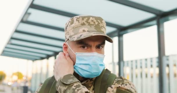 Porträtt av glad kaukasisk stilig ung manlig soldat i mössa tar av medicinsk mask på gatan. Närbild av självsäker man militarian utomhus i stan under pandemin. Militär uniform. — Stockvideo