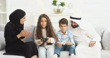 Küçük çocukları olan neşeli Arap ebeveynler kanepede oturup, joysticklerle video oyunu oynuyorlar. Anne ve babasıyla sevimli Arap çocukları evde kontrolörlerle video oyunları oynuyorlar..