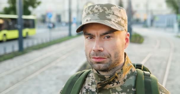 Porträtt av glad kaukasisk stilig ung manlig soldat i mössa med ryggsäck ser modig på gatan. Närbild av självsäker man militarian utomhus i stan. Militär uniform. — Stockvideo