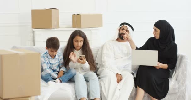 Arabští mladí rodiče v nádobí dasha a hidžáb sedí na gauči s notebookem a plánování interiéru pro nový domov po nastěhování. Malé děti mezi krabicemi hrají na smartphonu. Arabové s dětmi — Stock video