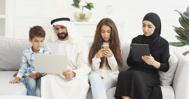 Arabian padre e hijo con ordenador portátil jugando en el ordenador. Madre e hija usando artilugios. Familia de Emiratos Árabes Unidos en casa. Padres con hijos. Dispositivos. Mujer musulmana con tableta. Chica golpeando en el teléfono inteligente. — Vídeos de Stock