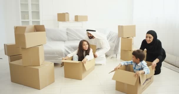 Vrolijke jonge moslims ouders in hijab en dishdasha spelen met kinderen in de woonkamer tijdens het verhuizen naar een nieuw huis. Arabieren moeder en vader rijden kleine zoon en dochter in kartonnen dozen. — Stockvideo