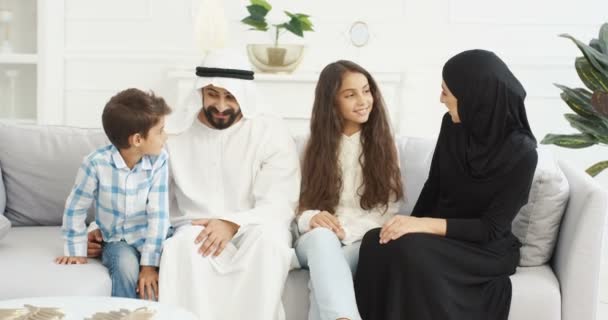 Retrato de família muçulmana alegre com dois filhos sentados no sofá e sorrindo alegremente para a câmera. Pequeno menino bonito e menina com mãe e pai em casa. Pais árabes com crianças pequenas abraçando . — Vídeo de Stock