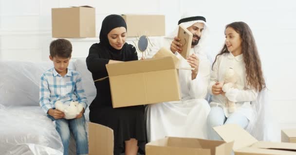 Χαρούμενοι Άραβες γονείς που κάθονται στον καναπέ στο σαλόνι με μικρά παιδιά και ξεπακετάρουν χαρτοκιβώτια. Μουσουλμανική οικογένεια με παιδιά που μετακομίζουν σε νέο σπίτι. Επισκευή. Ενήλικες και έφηβοι αλλάζουν κατοικία. — Αρχείο Βίντεο
