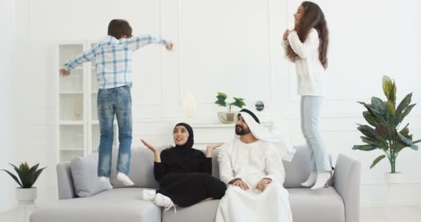 Madre y padre árabe predicando y calmando a los niños. Musulmanes padre y madre en ropa tradicional sentados en el sofá y sosteniendo la cabeza con las manos. Niños pequeños ruidosos activos saltando en el sofá jugando — Vídeo de stock