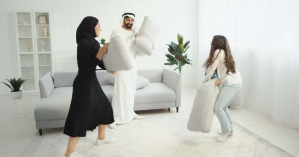 Arabische gelukkige vrolijke familie met kinderen die thuis spelen en plezier hebben. Wit interieur. Arabieren ouders en kinderen vechten met kussens in de woonkamer en lachen. Moslims in traditionele kleren. — Stockvideo