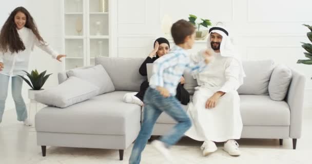 Два радостных арабских ребенка бегают кругами по гостиной. Счастливые симпатичные мальчик и девочка, играющие вокруг дивана. Улыбающиеся арабские мать и отец сидят на диване с ноутбуком дома — стоковое видео