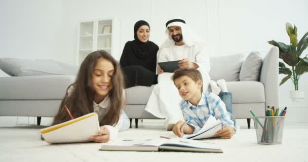 Des enfants arabes heureux assis sur le sol et peignant dans des cahiers. Petite sœur et frère dessinant à la maison. Deux parents assis sur le canapé et parlant. Arabes mère et père avec tablette dispositif. — Video