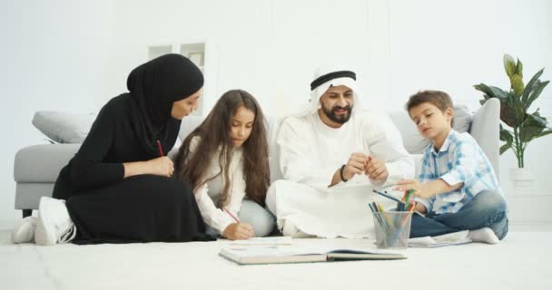 Arabska szczęśliwa rodzina z dziećmi siedzącymi na podłodze i malującymi w notatnikach. Dzieci z rodzicami rysującymi w domu. Arabowie rodzice w tradycyjnych ubraniach bawią się z synem i córką. — Wideo stockowe