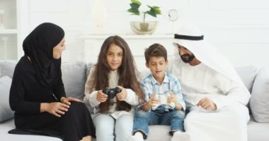 Mutlu neşeli Arap ailesi oturma odasında kanepede oturup joysticklerle video oyunu oynuyor. Müslüman anne ve babası olan sevimli çocuklar evde eğleniyor ve kontrolörlerle video oyunları oynuyorlar..