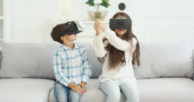 Beyaz tenli, VR gözlüklü şirin çocuklar oturma odasında beyaz koltukta oturuyorlar. Küçük tatlı bir kız ve oğlan, kız ve erkek kardeş evde oynuyor, sanal gerçeklik kulaklıkları var. Kapalı alanda. Geliştirilmiş oyun dünyası