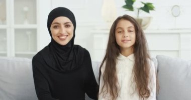 Oturma odasındaki kanepede tatlı genç kızıyla otururken kameraya gülümseyen başörtülü güzel Müslüman annenin portresi. Türbanlı güzel Arap kadın ve kanepedeki küçük genç kız..