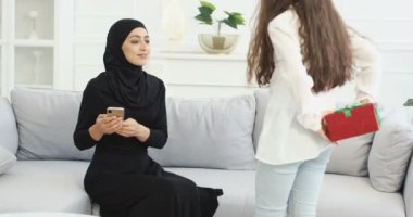 Kızı tesettürlü Müslüman bir anneye geliyor. Arkasında da hediye var. Abaya elbiseli Arap kadın koltukta oturuyor ve hediyesini kutuda teslim ediyor. Tatil, doğum günü ya da Anneler Günü hediyesi.