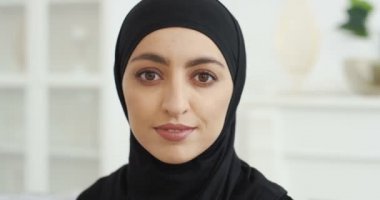 Siyah geleneksel tesettürlü güzel bir Arap kadının portresi doğrudan kameraya bakıyor ve gülümsüyor. - Evde. Gülümseyen güzel bir kadın yüzü. Arap başörtüsü. Kapalı.