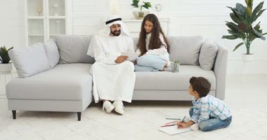 Beyaz kanduralı Arap bir baba oturma odasında küçük çocuklar, kız ve oğlanlarla oynuyor. Müslüman baba resim çiziyor ve boyuyor. Babasıyla evde resim yapan ve birlikte vakit geçiren bir kız ve erkek..
