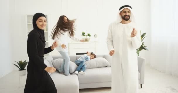 Fröhlich fröhliche arabische Mutter und Vater tanzen und amüsieren sich mit Tochter und Sohn im heimischen Wohnzimmer. Familientanz mit süßen Kindern und Lachen. Fröhliche Kinder genießen Musik mit den Eltern. — Stockvideo